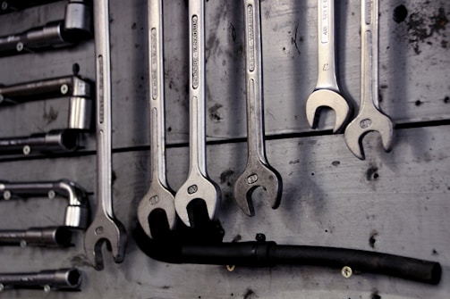 A collection of open-end and box-end wrenches hang on a weathered wooden board. The metal tools are neatly organized in a row, displaying their shiny metallic surfaces. A black hose or pipe with fittings is also visible below the wrenches.