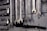 A collection of open-end and box-end wrenches hang on a weathered wooden board. The metal tools are neatly organized in a row, displaying their shiny metallic surfaces. A black hose or pipe with fittings is also visible below the wrenches.