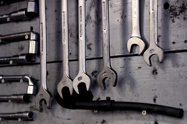 A collection of open-end and box-end wrenches hang on a weathered wooden board. The metal tools are neatly organized in a row, displaying their shiny metallic surfaces. A black hose or pipe with fittings is also visible below the wrenches.