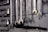 A collection of open-end and box-end wrenches hang on a weathered wooden board. The metal tools are neatly organized in a row, displaying their shiny metallic surfaces. A black hose or pipe with fittings is also visible below the wrenches.