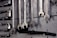 A collection of open-end and box-end wrenches hang on a weathered wooden board. The metal tools are neatly organized in a row, displaying their shiny metallic surfaces. A black hose or pipe with fittings is also visible below the wrenches.