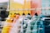 A row of colorful puffer jackets hangs on wooden hangers, displayed in a store window. The jackets vary in bright colors including yellow, orange, white, light blue, and dark blue. The background shows a reflection of the outside environment in the glass, adding a layered effect to the image.