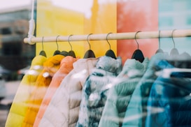 A row of colorful puffer jackets hangs on wooden hangers, displayed in a store window. The jackets vary in bright colors including yellow, orange, white, light blue, and dark blue. The background shows a reflection of the outside environment in the glass, adding a layered effect to the image.