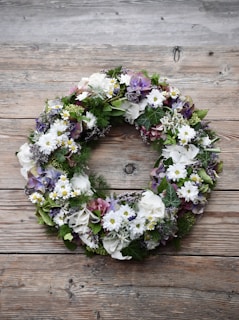 A beautifully crafted floral wreath consisting of white, purple, and green flowers and foliage arranged in a circular formation. The wreath is placed on a wooden surface with a rustic texture, enhancing the natural appeal. The flowers include daisies, hydrangeas, and various greenery.