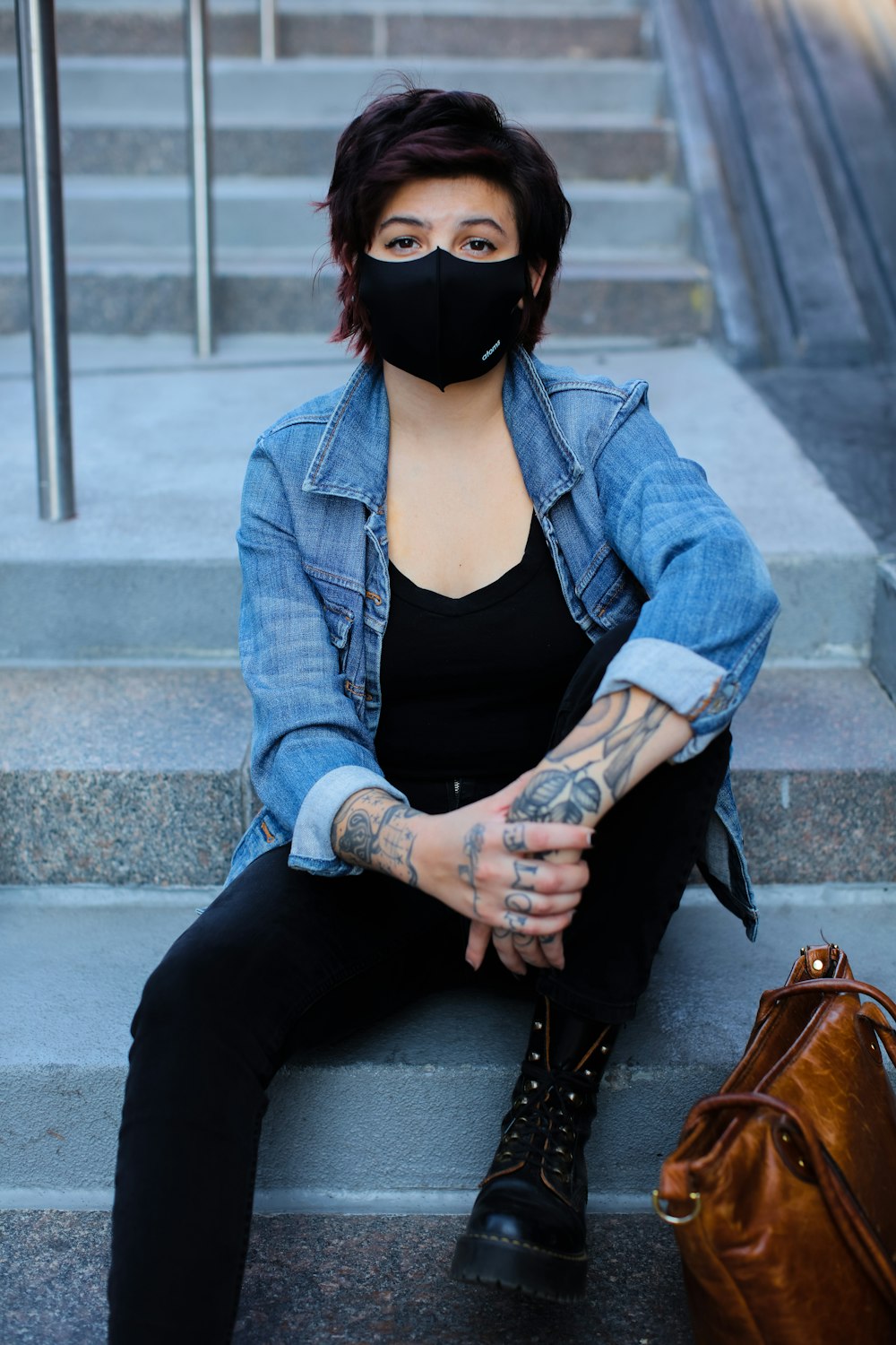 woman in blue denim jacket and black tank top wearing black mask sitting on concrete bench