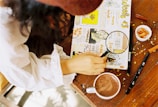 A person wearing a white shirt is holding a magnifying glass over a creatively decorated journal. The journal is filled with colorful illustrations, doodles, and handwritten text on a table. Nearby, there is a cup of cocoa, a pen, and a small bowl of snacks.