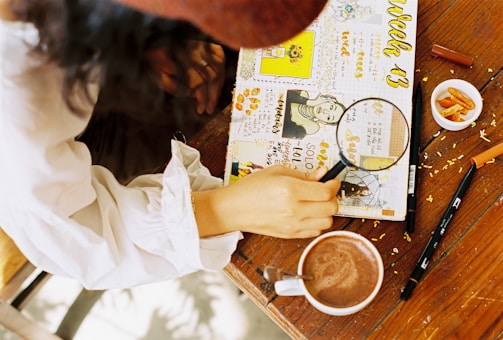 A person wearing a white shirt is holding a magnifying glass over a creatively decorated journal. The journal is filled with colorful illustrations, doodles, and handwritten text on a table. Nearby, there is a cup of cocoa, a pen, and a small bowl of snacks.