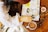 A person wearing a white shirt is holding a magnifying glass over a creatively decorated journal. The journal is filled with colorful illustrations, doodles, and handwritten text on a table. Nearby, there is a cup of cocoa, a pen, and a small bowl of snacks.
