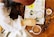 A person wearing a white shirt is holding a magnifying glass over a creatively decorated journal. The journal is filled with colorful illustrations, doodles, and handwritten text on a table. Nearby, there is a cup of cocoa, a pen, and a small bowl of snacks.