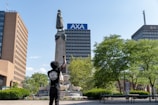 A person with raised fist stands in front of a tall monument featuring a statue. Surrounding the monument are lush green trees, and in the background are several tall buildings, including one with the 'AXA' logo. The person is wearing a black t-shirt with a graphic on the back.