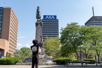 A person with raised fist stands in front of a tall monument featuring a statue. Surrounding the monument are lush green trees, and in the background are several tall buildings, including one with the 'AXA' logo. The person is wearing a black t-shirt with a graphic on the back.