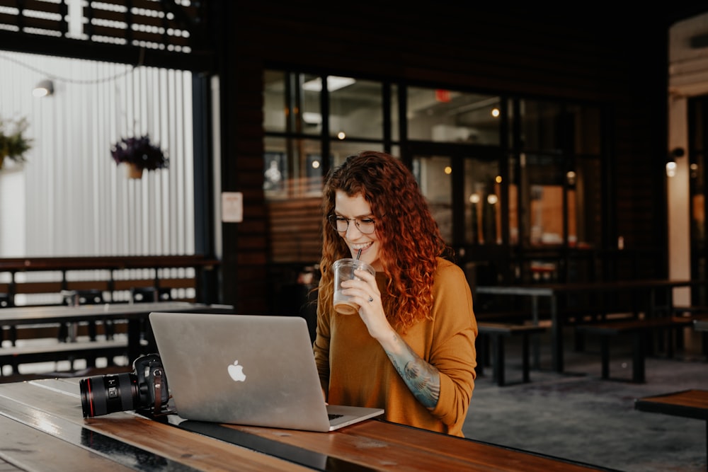 curly ginger woman in orange long sleeve shirt sitting in front of silver macbook sipping iced coffee smiling in an empty coffee shop