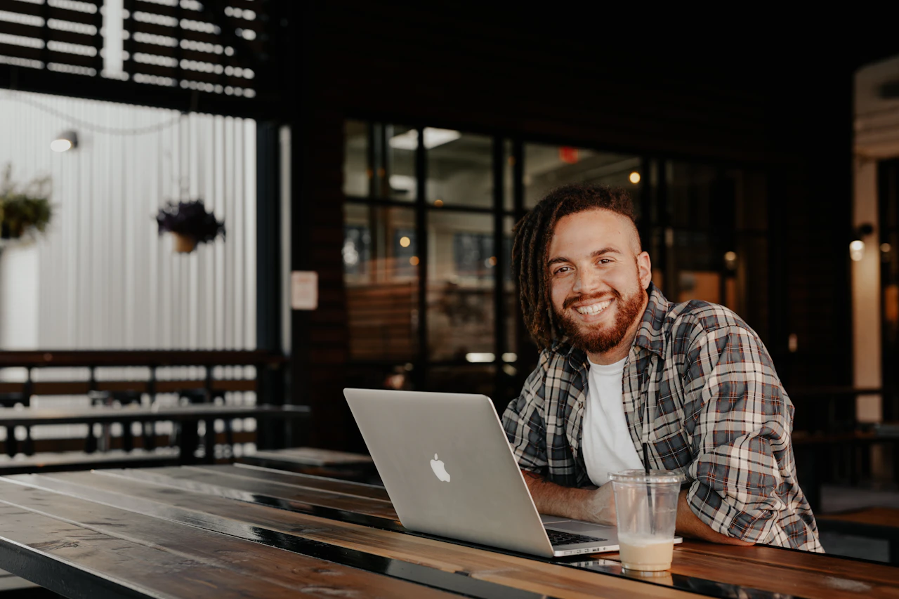 Smiling man with a laptop sits alone at a long wooden table in a quiet, empty café.