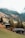 A serene mountain landscape features wooden cabins nestled on a grassy hillside. Dense evergreen forests cover the slopes in the background, with fog drifting among the trees. The scene exudes a peaceful and idyllic rural ambiance.