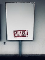 A large rectangular blank billboard mounted on a wall with the text 'KULTUR AFFOLTERN' in bold red letters near the bottom center. The billboard is framed with a metallic border and is positioned in an outdoor setting against a textured, light-colored wall.