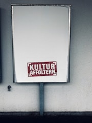 A large rectangular blank billboard mounted on a wall with the text 'KULTUR AFFOLTERN' in bold red letters near the bottom center. The billboard is framed with a metallic border and is positioned in an outdoor setting against a textured, light-colored wall.