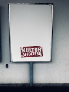 A large rectangular blank billboard mounted on a wall with the text 'KULTUR AFFOLTERN' in bold red letters near the bottom center. The billboard is framed with a metallic border and is positioned in an outdoor setting against a textured, light-colored wall.