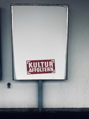 A large rectangular blank billboard mounted on a wall with the text 'KULTUR AFFOLTERN' in bold red letters near the bottom center. The billboard is framed with a metallic border and is positioned in an outdoor setting against a textured, light-colored wall.
