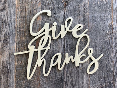 A decorative script of the words 'Give Thanks' is displayed in a light-colored, cursive font. It is set against a background of rustic, weathered wood with a grayish-brown tone.
