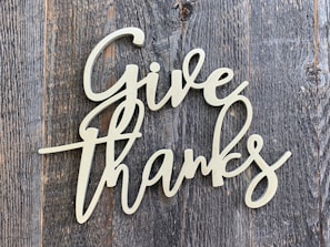 A decorative script of the words 'Give Thanks' is displayed in a light-colored, cursive font. It is set against a background of rustic, weathered wood with a grayish-brown tone.