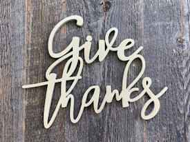 A decorative script of the words 'Give Thanks' is displayed in a light-colored, cursive font. It is set against a background of rustic, weathered wood with a grayish-brown tone.