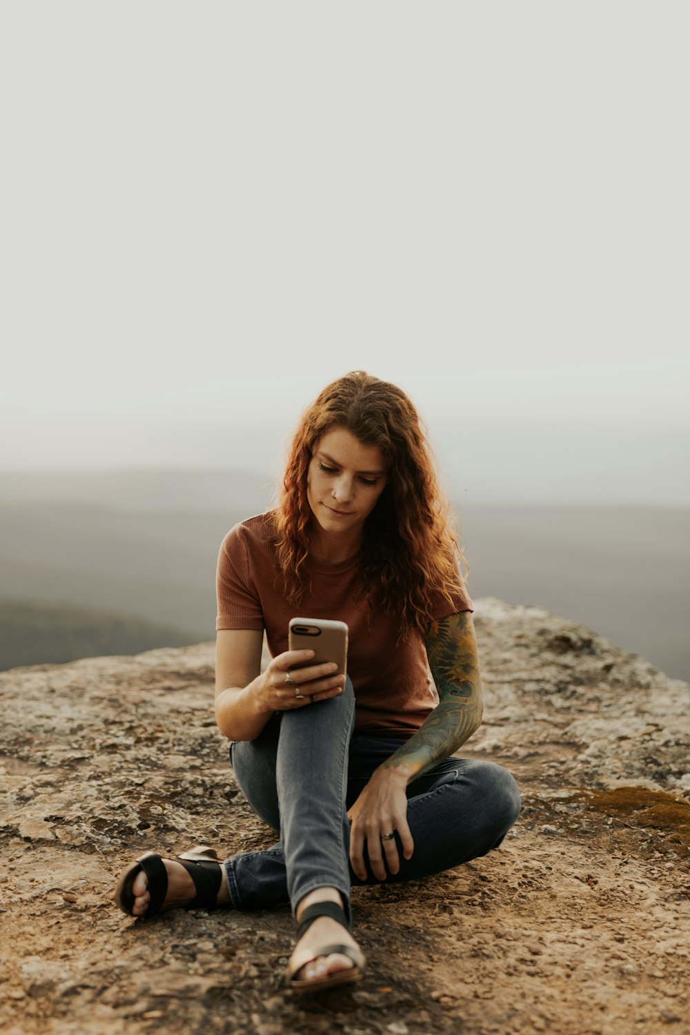 woman in brown long sleeve shirt and blue denim jeans sitting on rock during daytime
