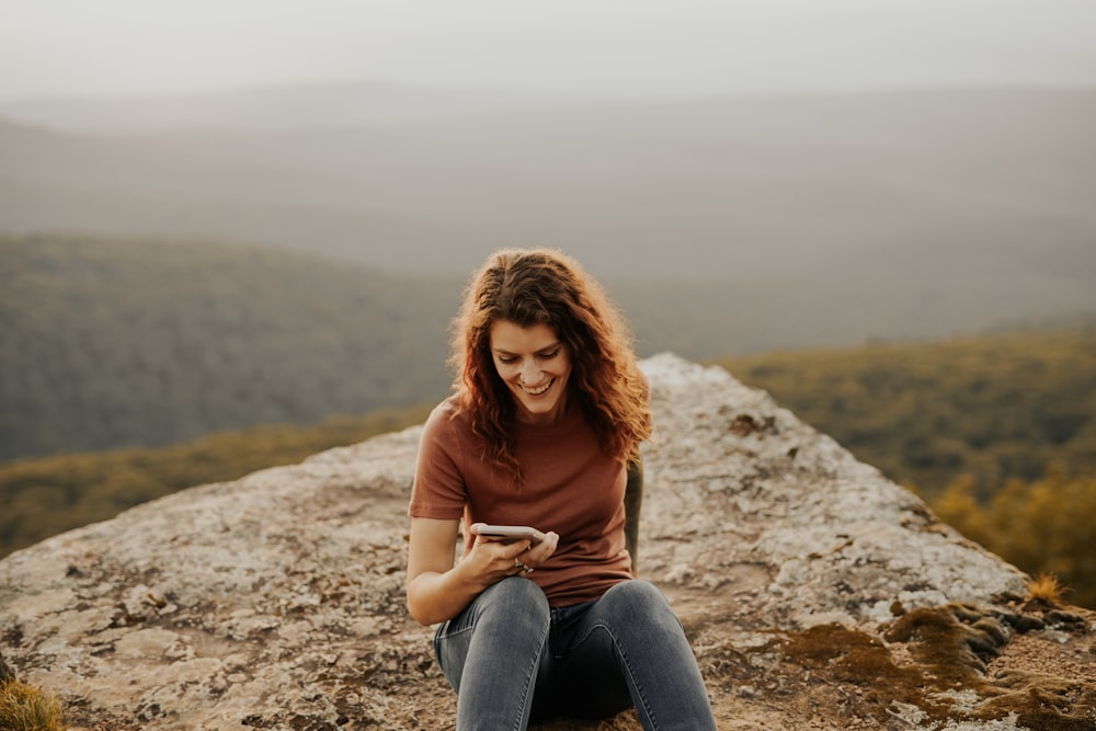 woman in red long sleeve shirt and blue denim jeans sitting on rock during daytime