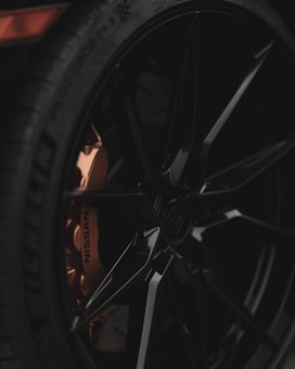 Close-up view of a black car wheel with a shiny, sleek design. The visible portion of the brake caliper is copper-colored with the word 'NISSAN' imprinted on it. The tire brand 'MICHELIN' is also visible on the side of the tire.