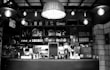 A cozy café interior with various kitchen appliances and utensils neatly arranged on shelves behind a counter. The counter features coffee machines, teapots, and a variety of jars and glassware. The space is warmly lit with hanging industrial-style lights and features wooden and brick elements.