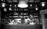 A cozy café interior with various kitchen appliances and utensils neatly arranged on shelves behind a counter. The counter features coffee machines, teapots, and a variety of jars and glassware. The space is warmly lit with hanging industrial-style lights and features wooden and brick elements.