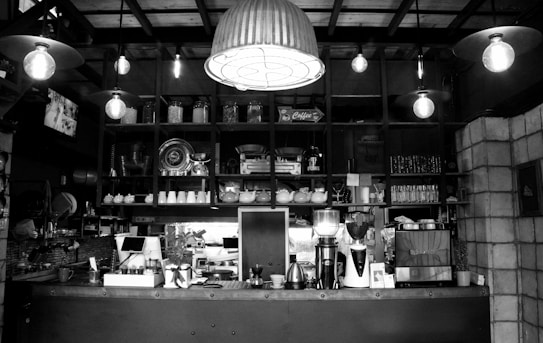 A cozy caf&eacute; interior with various kitchen appliances and utensils neatly arranged on shelves behind a counter. The counter features coffee machines, teapots, and a variety of jars and glassware. The space is warmly lit with hanging industrial-style lights and features wooden and brick elements.