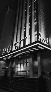 Neon-lit building facade with a large 'POST' sign glows in the night. The entrance is lined with patterned tiles and glass doors. Two people holding umbrellas walk past the building on a rainy evening, casting soft shadows on the ground.
