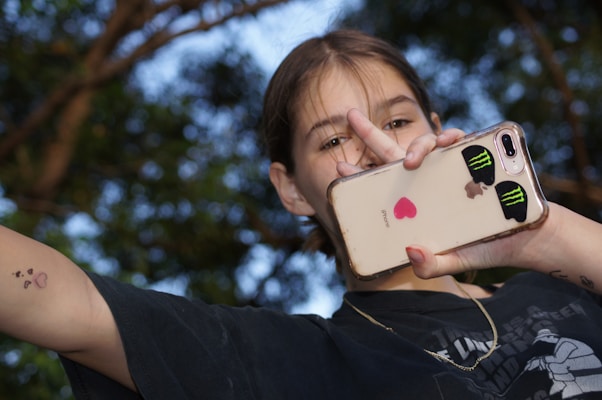A person holding a smartphone with decorative stickers, including a pink heart and two green Monster Energy logos. The person is wearing a black t-shirt and a necklace, and they are making a peace sign with their fingers. The background includes tree branches with a natural, blurred effect.