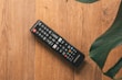 A television remote control rests on a wooden surface. The remote is black with various colored buttons and has a brand label. Large green leaves partially frame the image from the top right corner.