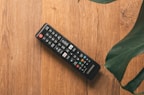 A television remote control rests on a wooden surface. The remote is black with various colored buttons and has a brand label. Large green leaves partially frame the image from the top right corner.
