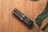 A television remote control rests on a wooden surface. The remote is black with various colored buttons and has a brand label. Large green leaves partially frame the image from the top right corner.