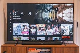 A television screen displaying the Netflix interface is shown. The main focus is the show 'Dark' with a large image from the series. Below it are recommendations for 'Continue Watching,' including shows like 'Selling Sunset,' 'Friends,' and 'Community.' The interface includes the Netflix logo and an array of show thumbnails.
