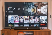 A television screen displaying the Netflix interface is shown. The main focus is the show 'Dark' with a large image from the series. Below it are recommendations for 'Continue Watching,' including shows like 'Selling Sunset,' 'Friends,' and 'Community.' The interface includes the Netflix logo and an array of show thumbnails.