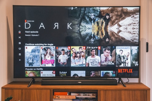 A television screen displaying the Netflix interface is shown. The main focus is the show 'Dark' with a large image from the series. Below it are recommendations for 'Continue Watching,' including shows like 'Selling Sunset,' 'Friends,' and 'Community.' The interface includes the Netflix logo and an array of show thumbnails.