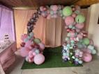 An elegantly decorated area featuring a balloon arch made up of pastel pink, green, and purple balloons adorned with small white butterflies. The backdrop curtain is pale pink with a row of artificial flowers intermixed with the balloons. On the left is a table with cake pops and cupcakes, adding to the festive atmosphere. The scene conveys a sense of preparation for a party or celebration in a cozy, indoor setting.