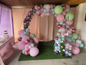 An elegantly decorated area featuring a balloon arch made up of pastel pink, green, and purple balloons adorned with small white butterflies. The backdrop curtain is pale pink with a row of artificial flowers intermixed with the balloons. On the left is a table with cake pops and cupcakes, adding to the festive atmosphere. The scene conveys a sense of preparation for a party or celebration in a cozy, indoor setting.