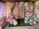 An elegantly decorated area featuring a balloon arch made up of pastel pink, green, and purple balloons adorned with small white butterflies. The backdrop curtain is pale pink with a row of artificial flowers intermixed with the balloons. On the left is a table with cake pops and cupcakes, adding to the festive atmosphere. The scene conveys a sense of preparation for a party or celebration in a cozy, indoor setting.