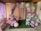 An elegantly decorated area featuring a balloon arch made up of pastel pink, green, and purple balloons adorned with small white butterflies. The backdrop curtain is pale pink with a row of artificial flowers intermixed with the balloons. On the left is a table with cake pops and cupcakes, adding to the festive atmosphere. The scene conveys a sense of preparation for a party or celebration in a cozy, indoor setting.