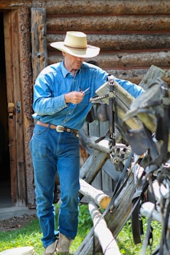 A person wearing a blue denim shirt and jeans, along with a straw hat, is seen outside a rustic wooden structure. They are holding a tool and interacting with horse tack or a similar item hung on a wooden fence.