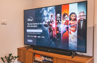 A television displaying a Disney+ promotional screen featuring various characters from popular franchises. Below the screen is a wooden cabinet with a few books and a small plant. The wall is light-colored, creating a cozy and inviting atmosphere.