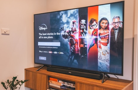 A television displaying a Disney+ promotional screen featuring various characters from popular franchises. Below the screen is a wooden cabinet with a few books and a small plant. The wall is light-colored, creating a cozy and inviting atmosphere.
