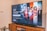 A television displaying a Disney+ promotional screen featuring various characters from popular franchises. Below the screen is a wooden cabinet with a few books and a small plant. The wall is light-colored, creating a cozy and inviting atmosphere.