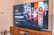 A television displaying a Disney+ promotional screen featuring various characters from popular franchises. Below the screen is a wooden cabinet with a few books and a small plant. The wall is light-colored, creating a cozy and inviting atmosphere.