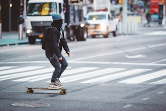 A person wearing a beanie and a face covering rides a skateboard across a city street. The individual is dressed in casual attire, including a backpack, as they navigate on a longboard in an urban setting. Vehicles are visible in the background, emphasizing the busy city environment.