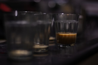 Several empty and partially filled glass tumblers are lined up on a dark surface. The focus is on a single glass containing a small amount of a brown liquid, possibly coffee. The background and other glasses are blurred, suggesting a dimly lit environment and an emphasis on the glass with liquid.